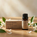 A small amber glass bottle of Neroli Mystica Immaculate Joy essential oil blend stands next to its kraft paper box on a wooden surface, surrounded by white neroli blossoms and green leaves.