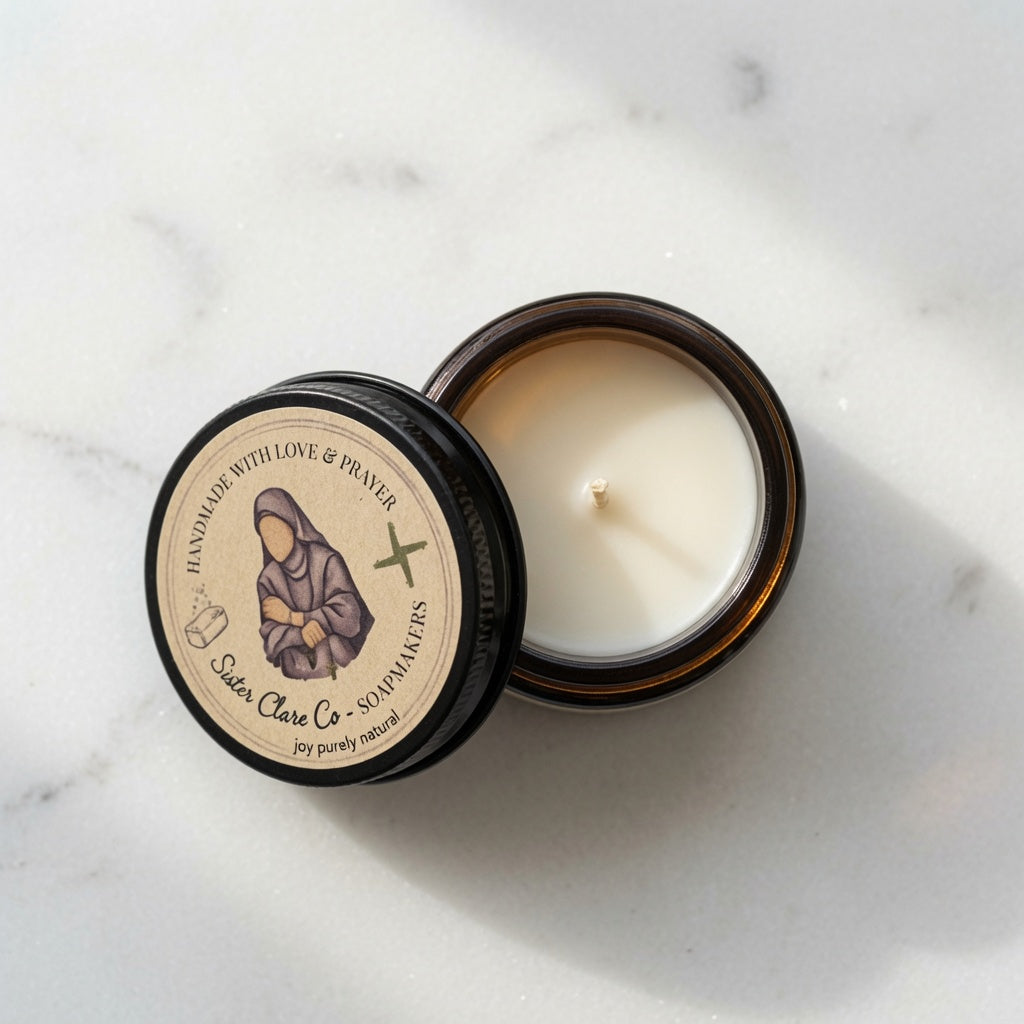 An open amber glass jar candle with white wax sits on a light marble surface next to its lid. The lid features a beige label with an illustration of a robed figure and the text 'Sisters Clare Co - St.