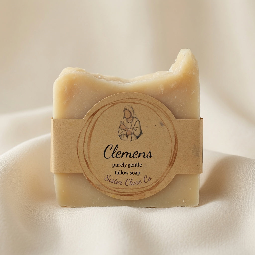 A bar of Clemens tallow soap wrapped in a kraft paper band with a round label, set against a soft, draped cream fabric background.
