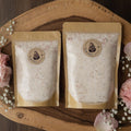 Square product photograph of small and large Athleta Christi Mineral Soak pouches arranged side by side on natural wood or stone surface with soft shadows, floral accents, and elegant styling that helps customers visualize stocking up for their faith-filled recovery routine.