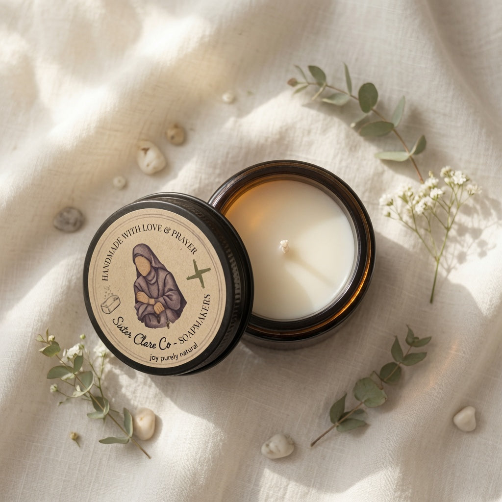 An open amber glass candle jar with cream-colored wax sits on a textured cream linen fabric, surrounded by small white stones and sprigs of eucalyptus. The lid is placed beside the jar, showing a round beige label with a dark illustration and the text 'Scent of Life Co - Soul Smakers'.