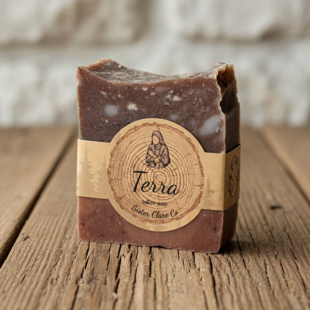 Hero shot of Terra Tallow Soap bar standing upright on a warm rustic wooden surface, highlighting its layered earth-crust colors, marbling, rough edges, matte finish, and Sister Clare Co. circular nun logo label in crisp product-photography style.